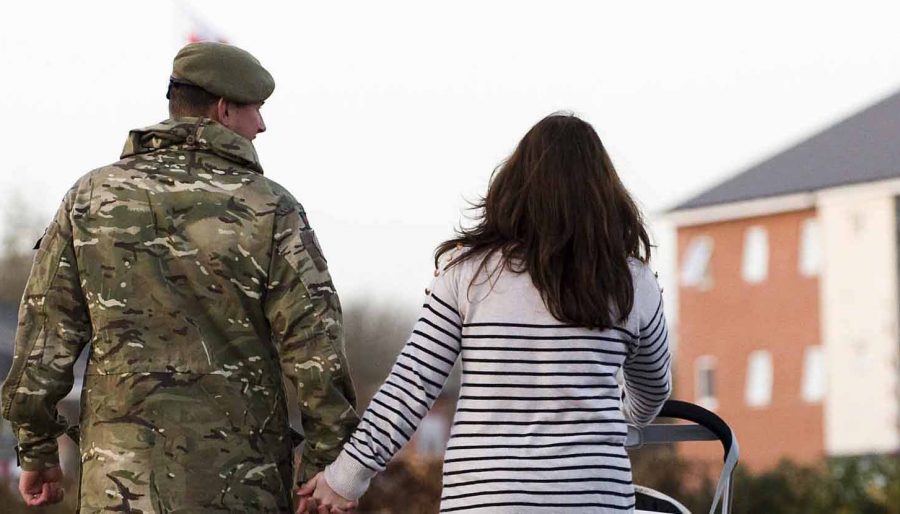 Image showing a soldier returning home, holding hands with his wife. She is pushing a stroller in front of buildings in the background.