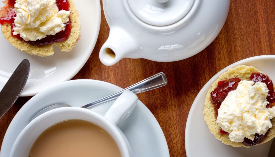 Image showing a pot and cup of tea with two scones with jam and cream. There is also cutlery and plates and saucers.