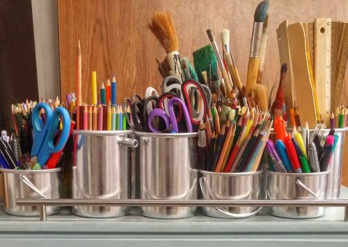 Image of various arts and crafts utensils in pots. There are scissors, paint brushes, pens, rulers and colouring pencils.